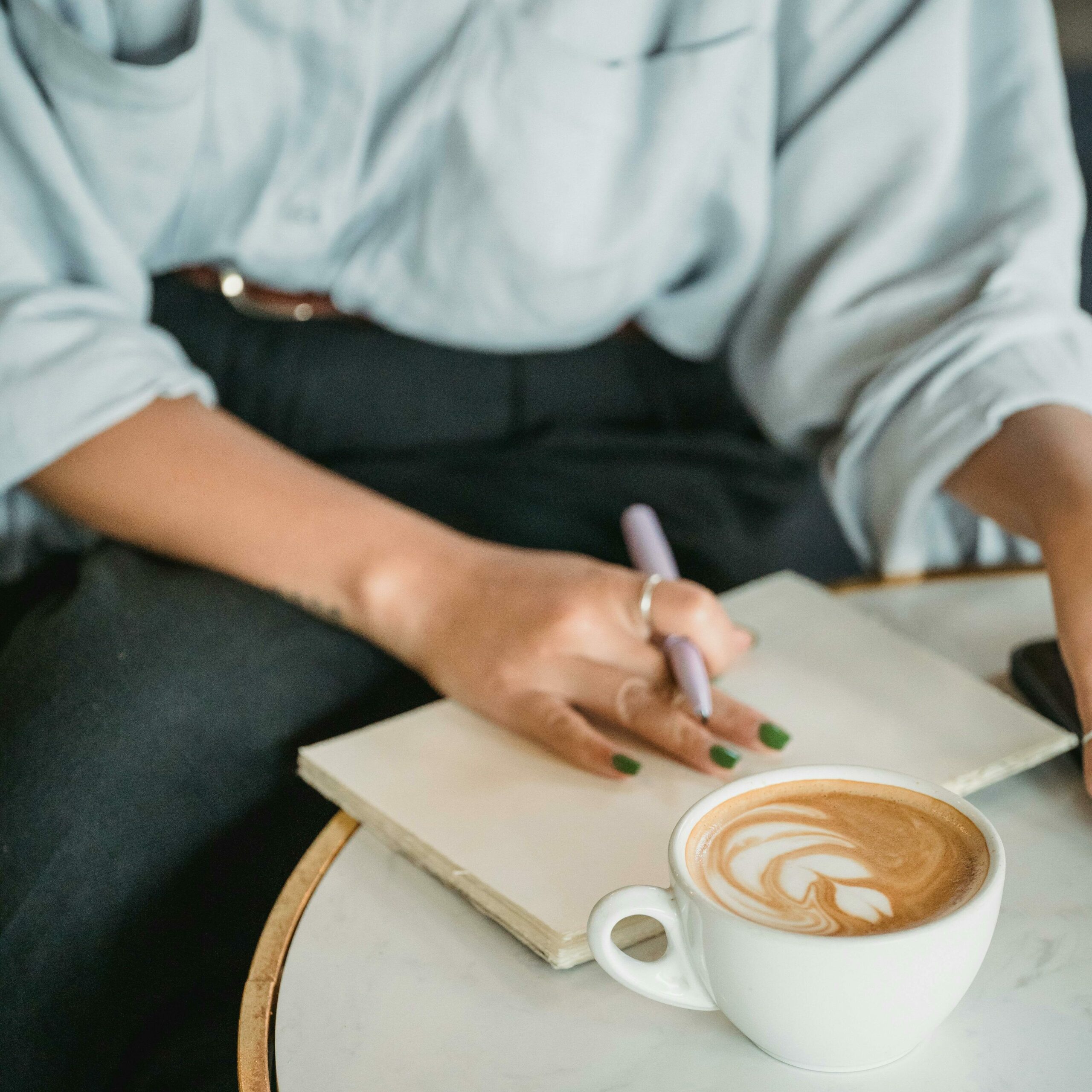 Une femme tient un stylo et pose sa main sur un carnet. A côté d'elle se trouve une tasse de café. Cela illustre le fait les entretiens se déroulent dans le lieu choisi par le biographé.